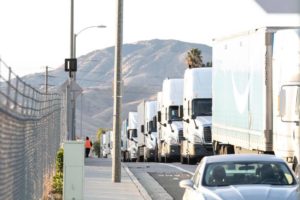 A line of large semi-trucks is parked along the side of a road with a chain-link fence on the left. The trucks are positioned one after another. In the background, there are hills and a few scattered palm trees, evoking scenes often captured by Berkeley Journalism students. A car is parked on the right side of the road.