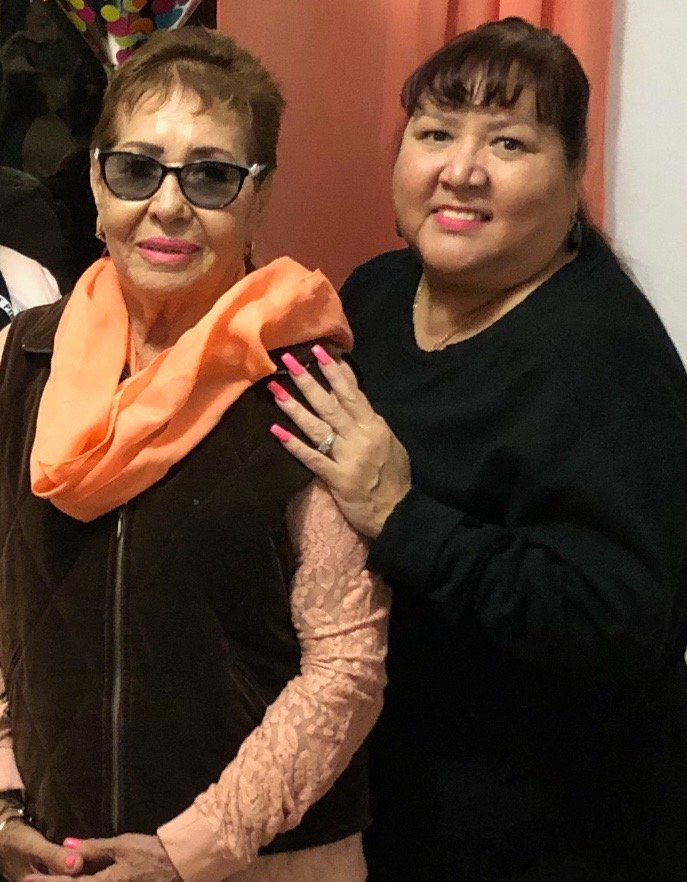 Two smiling women pose together against a light background. The woman on the left, wearing sunglasses, an orange scarf, and a brown jacket, seems to exude Berkeley Journalism chic. The woman on the right, resting her arm on the other's shoulder, wears a black top and has long, dark hair.