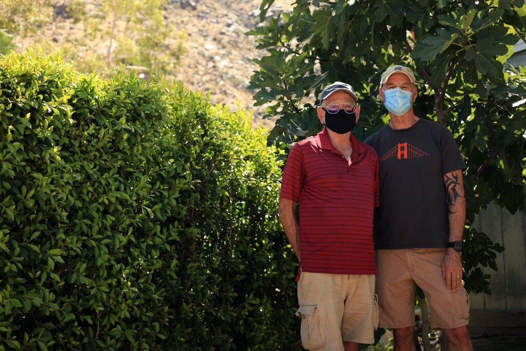 Two men wearing face masks stand outdoors in front of tall, leafy bushes. Both are dressed casually in shorts; the man on the left sports a red striped shirt, while the man on the right wears a black T-shirt and cap. They pose under a tree in sunny weather, perhaps discussing Berkeley Journalism.