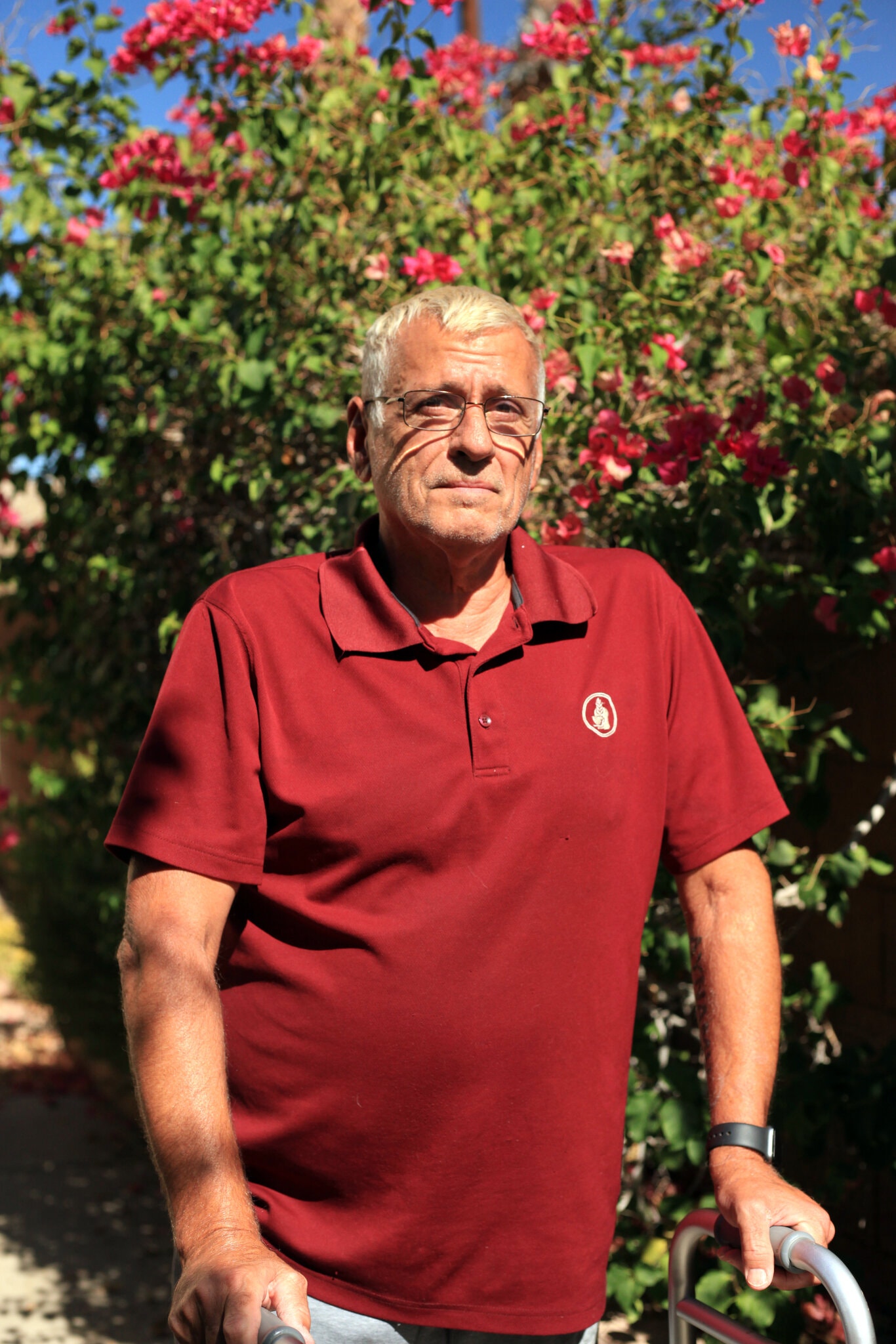 An elderly man with short white hair and glasses stands outdoors in front of a lush, flowering bush. Wearing a maroon polo shirt and using a walker, he seems deep in thought. The sun shines brightly, casting clear shadows and enhancing the colors of the flowers—capturing a scene worthy of Berkeley Journalism.
