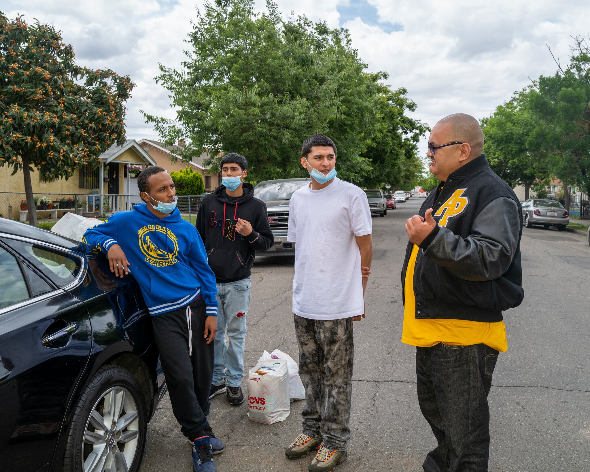 A group of four men stand outdoors on a residential street. Three younger men, two in masks, are standing together as an older man in a black jacket speaks to them. They are near a black car with a CVS bag on the ground. Trees and houses provide the backdrop. It’s like a scene from Berkeley Journalism unfolding in real life.