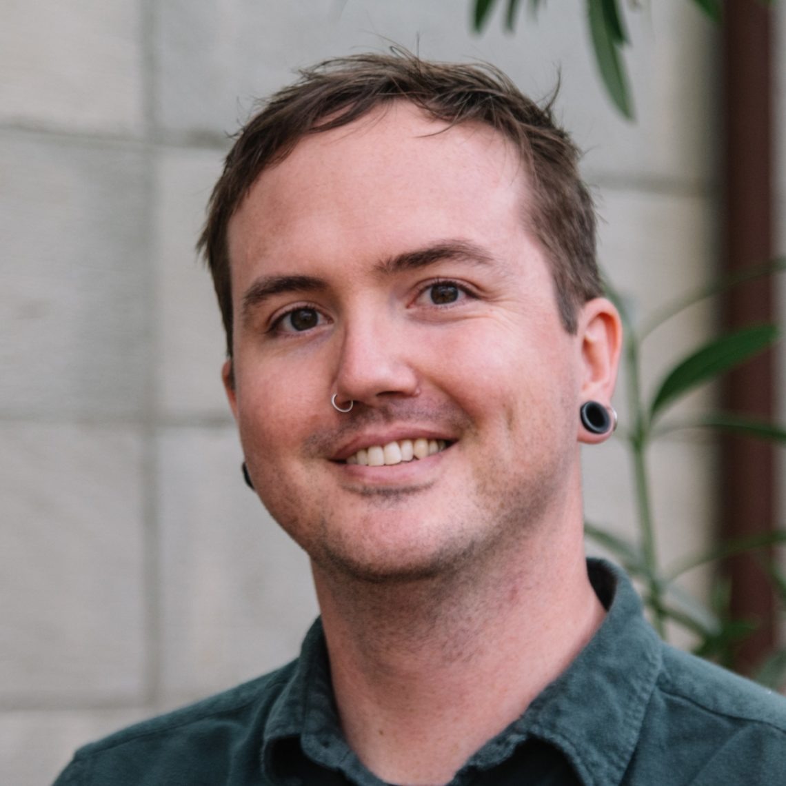A person with short brown hair, a nose piercing, and gauged earrings smiles while facing the camera. They are wearing a dark green button-up shirt with a background of light-colored stone walls and green foliage, capturing the essence of Berkeley Journalism's vibrant community spirit.