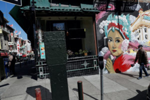Street scene in Chinatown featuring a detailed mural of a woman in traditional Chinese opera attire on a building. A man walks by the mural, and several pedestrians, possibly students from Berkeley Journalism, are seen in the background. Storefronts and street signage add to the urban atmosphere.