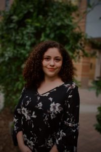 A person with curly hair, wearing a black floral blouse, stands outdoors in front of green foliage and a blurred building. They are smiling and looking at the camera, embodying the vibrant spirit of Berkeley Journalism.