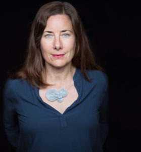 A woman with shoulder-length brown hair and light eyes is standing against a dark background. She is wearing a blue long-sleeve top with a distinctive, circular silver necklace. With a neutral expression, she looks directly at the camera, embodying the poised professionalism often seen in Berkeley Journalism graduates.
