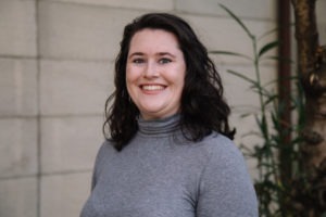 A person with shoulder-length dark hair, wearing a gray turtleneck sweater, smiles while standing in front of a light-colored wall and some greenery, embodying the optimism and energy often seen at Berkeley Journalism.