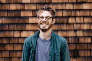A man with glasses and a beard, who we believe is Ben Derico, smiles at the camera. He is wearing a green shirt over a gray t-shirt. The background is a wall covered with wooden shingles.