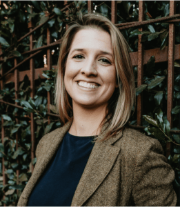 A person with shoulder-length hair wearing a blazer and a dark shirt stands smiling in front of a leafy background with a metal fence partially visible, embodying the vibrant spirit of Berkeley Journalism.