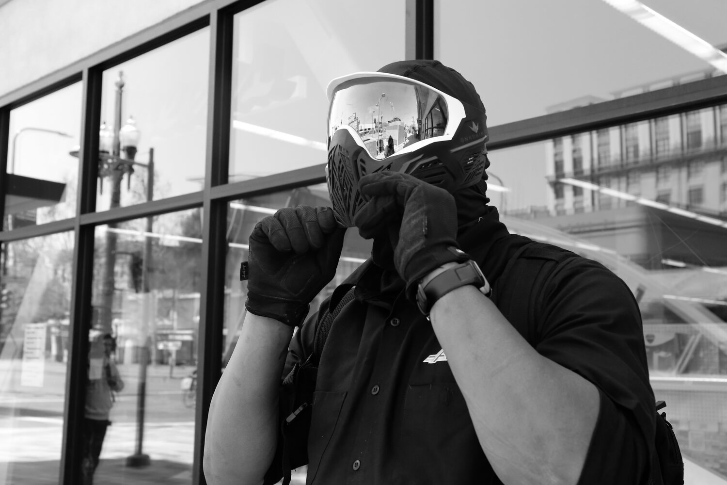 A person wearing a mask, goggles, and gloves adjusts their face covering outdoors. Their reflection and surrounding buildings are visible in the mirrored goggles. Standing near a glass building with street lamps and other urban elements in the background, they evoke a scene straight out of Berkeley Journalism.