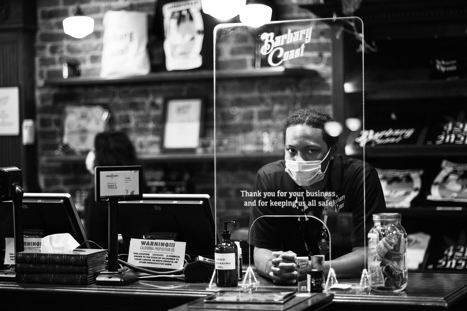 Black and white photo of a store employee wearing a face mask, standing behind a glass partition with text: 