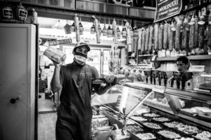 A worker in a deli shop wearing a face mask holds packaged meats and gestures with his right hand. Behind him, shelves are stocked with hanging sausages, and a glass display case filled with various food items is visible in the foreground. Another person works in the background, capturing scenes for Berkeley Journalism.