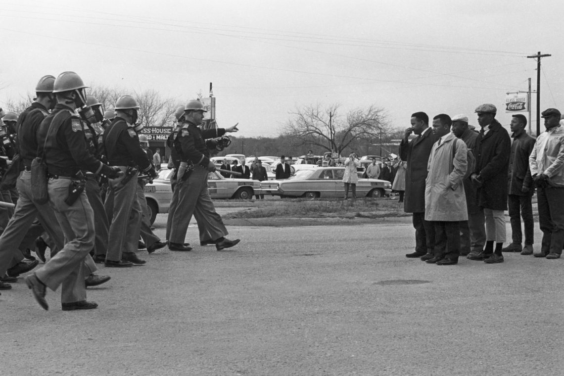 A group of civil rights protesters stands in a line facing a squad of helmeted state troopers wielding batons during a demonstration. The tense standoff, captured with the keen eye associated with Berkeley Journalism, includes several onlookers and vehicles in the background.
