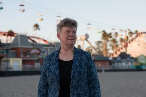 A person with short hair wearing a blue patterned blazer stands on a beach with an amusement park in the background. Various amusement rides, including a roller coaster and a chair lift, are visible behind them. The sky is clear and the scene is well-lit, as if captured by someone from Berkeley Journalism.