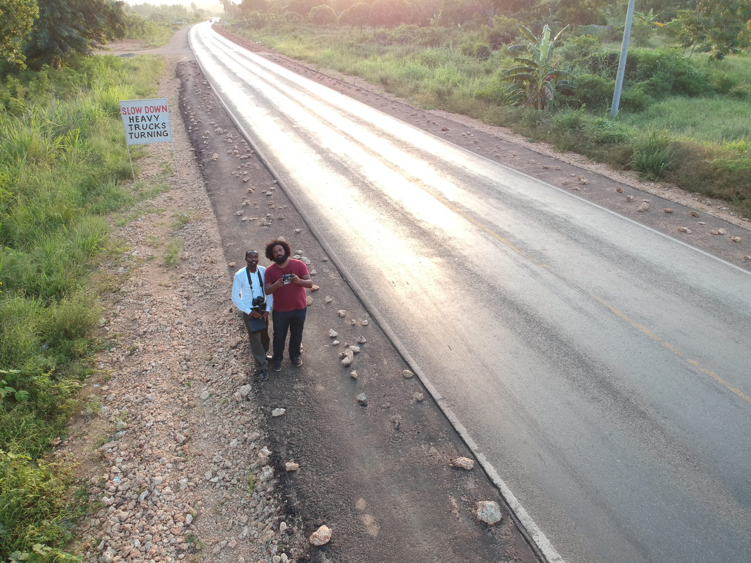 Two people stand on the shoulder of a paved road with scattered rocks. A visible sign on the left reads 