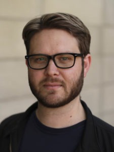 A man with short brown hair, a beard, and glasses is looking at the camera. He is wearing a black jacket over a dark shirt, resembling someone who might be entrenched in Berkeley Journalism. The background is blurred, giving the impression of an outdoor setting.