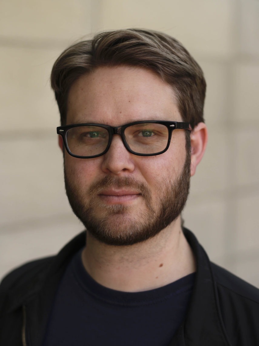 A close up headshot of a young man with brown short hair, glasses, and a beard.