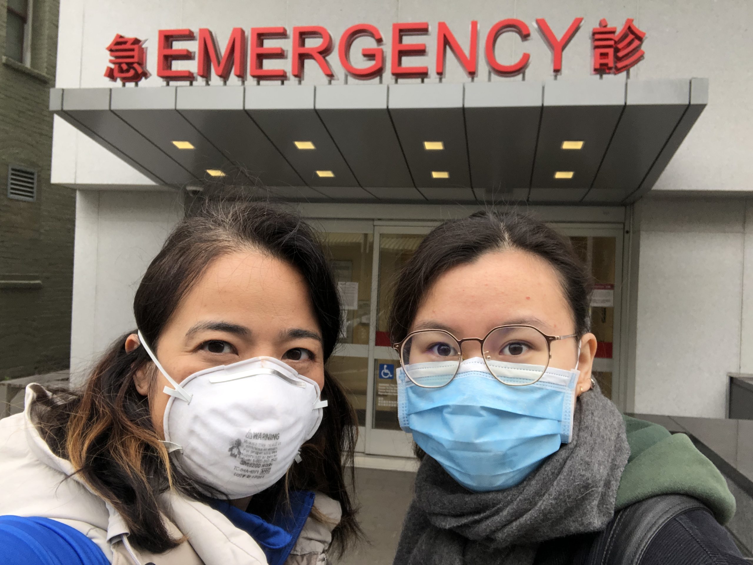 Two individuals wearing face masks stand in front of an emergency room entrance. The emergency sign is prominently displayed above them in both English and a second language. The atmosphere appears serious as they look toward the camera, capturing a scene reminiscent of a hard-hitting Berkeley Journalism report.