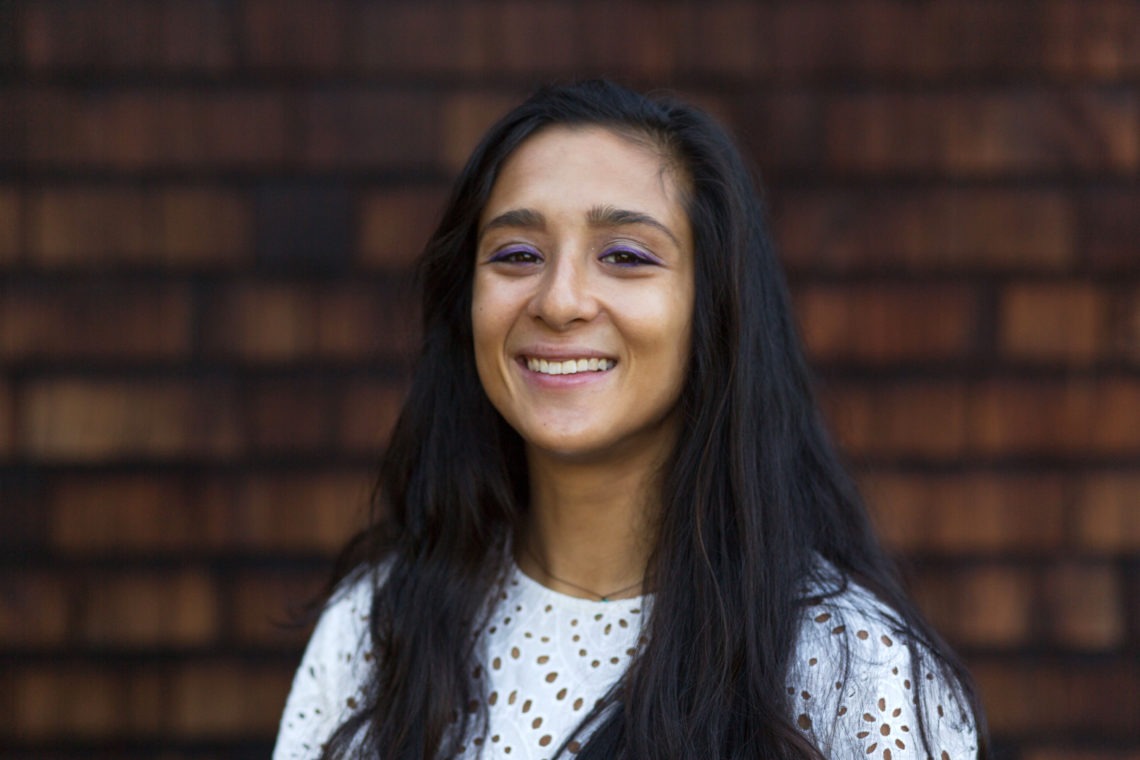A person with long black hair and light purple eyeshadow smiles at the camera. They are wearing a white top with eyelet details and are standing in front of a wooden shingle background, capturing the essence of Berkeley Journalism.