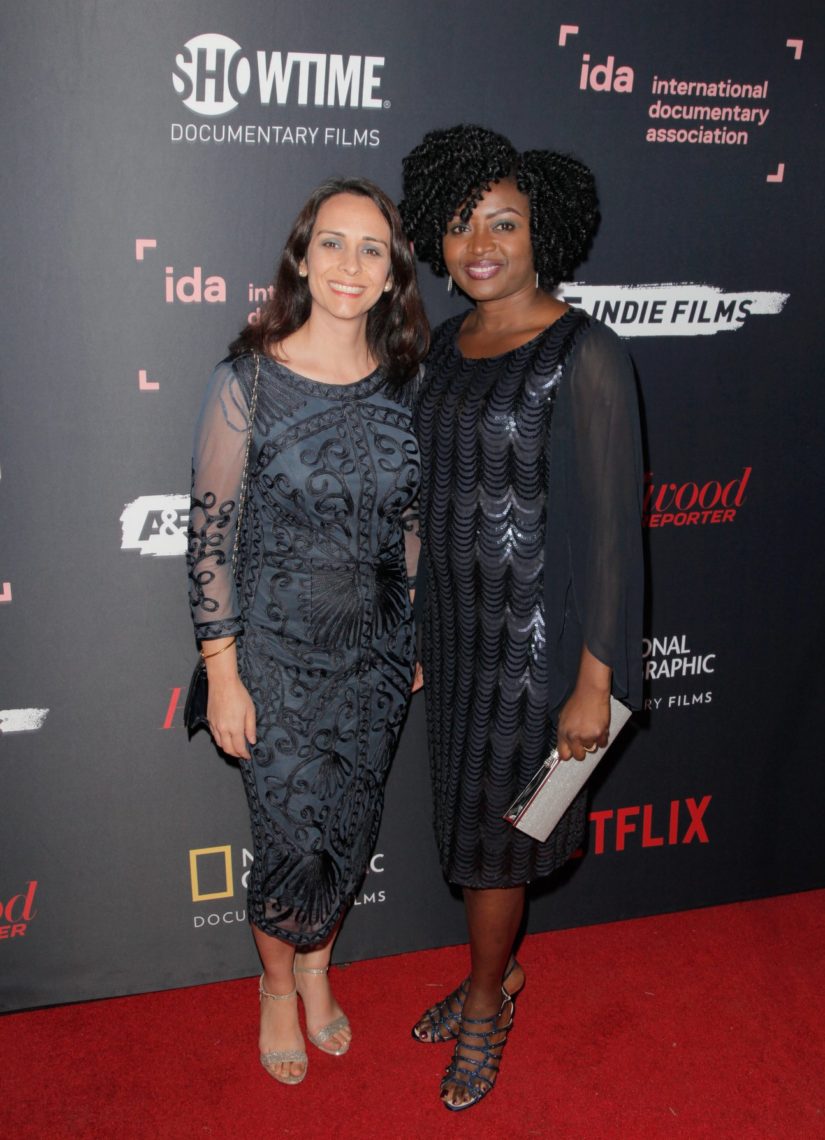 Two women, graduates of Berkeley Journalism, stand together on the red carpet, smiling for the camera. Both are dressed in elegant dresses; the woman on the left is in navy blue, while the woman on the right wears a black dress with wavy patterns. Various sponsor logos frame their picture-perfect moment.