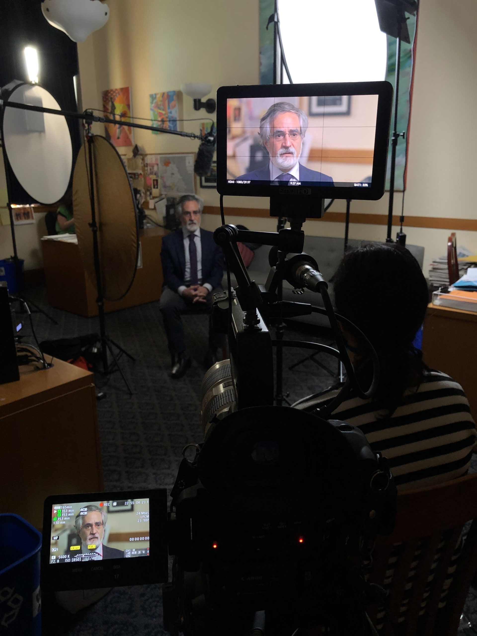 A behind-the-scenes look at a video interview setup in an office. A man in a suit sits in a chair, facing the camera. Professional lighting and filming equipment surround him, with camera monitors displaying close-up shots of the interviewee. This setup captures the high standards of Berkeley Journalism.
