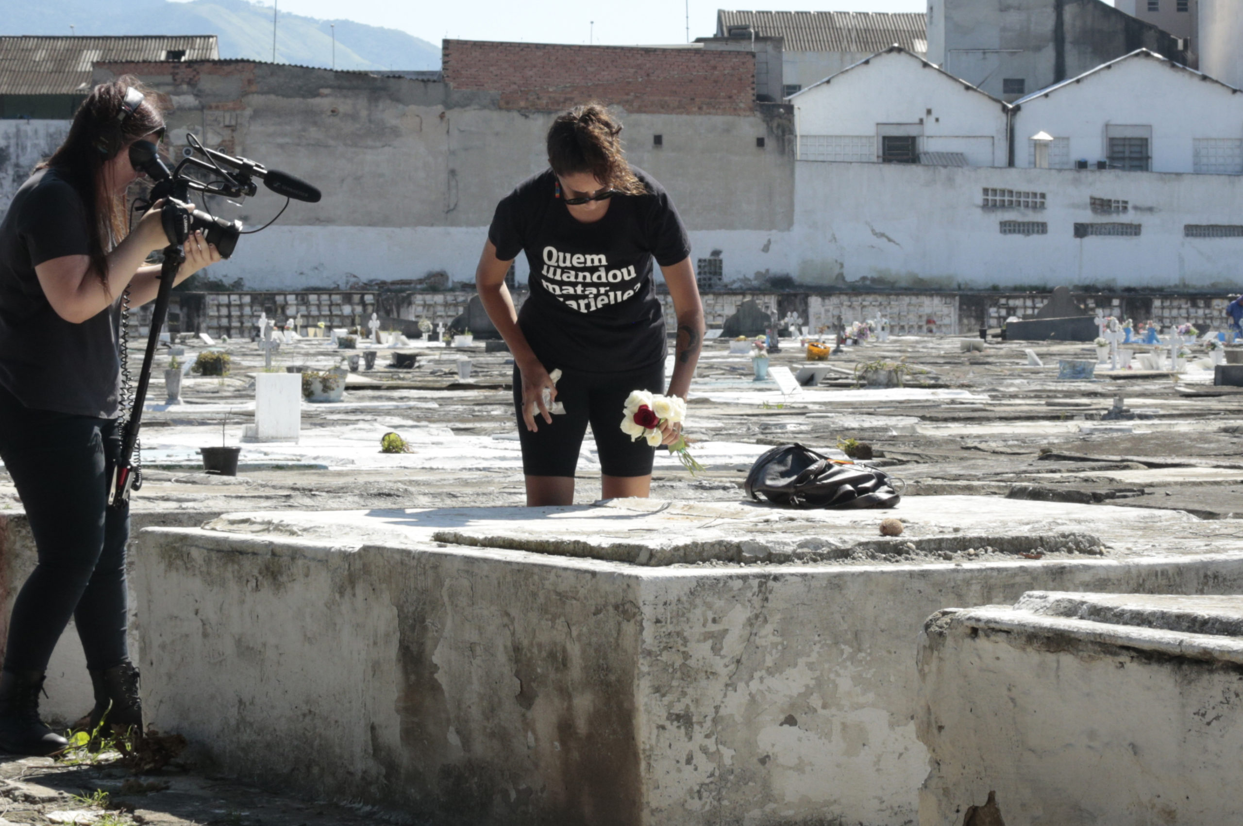 Two people in a cemetery appear to be filming. One holds a camera while the other, holding a bouquet of flowers, stands near a grave, wearing a T-shirt with Portuguese text. In the background are various gravestones and buildings—a potential project by Berkeley Journalism students capturing poignant moments.