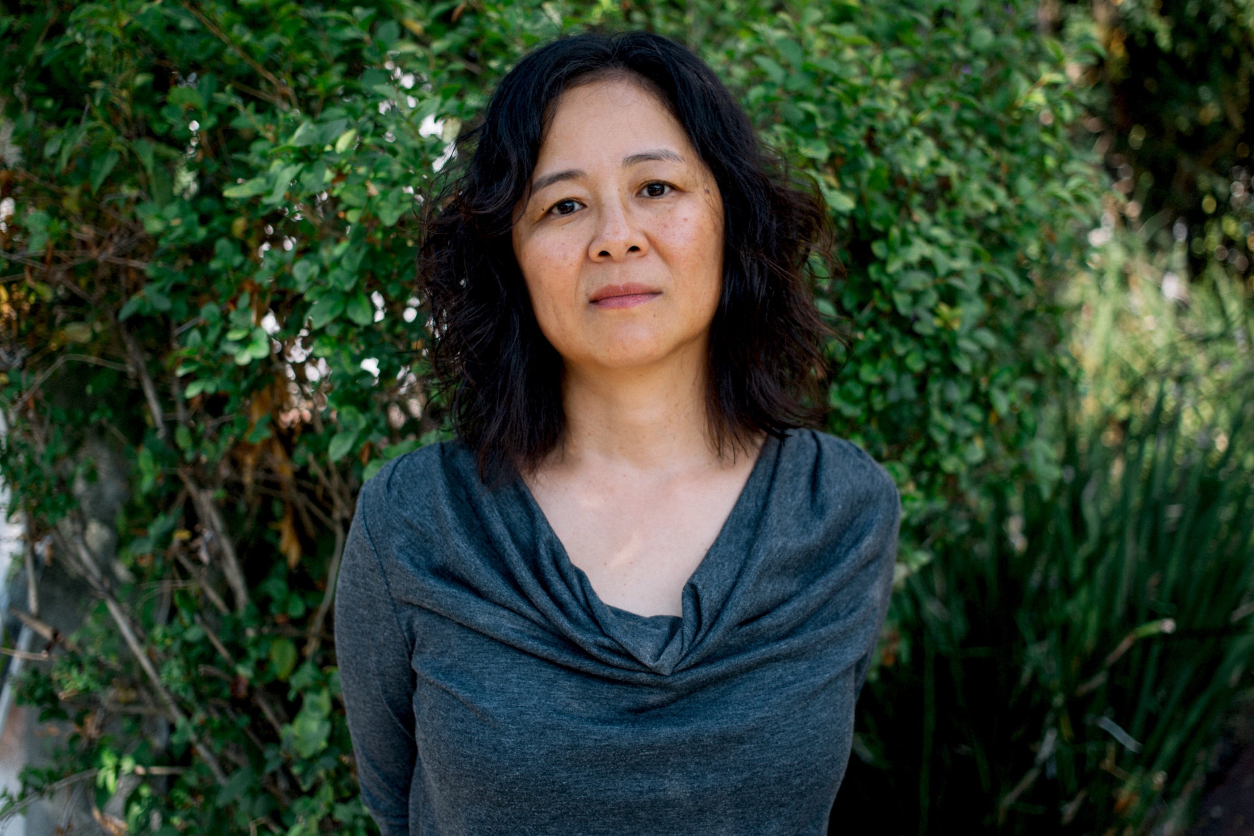 A woman with shoulder-length dark hair and a serious expression stands outdoors in front of lush green foliage. She is wearing a charcoal gray cowl neck top. The background, reminiscent of a Berkeley Journalism setting, is filled with dense greenery.