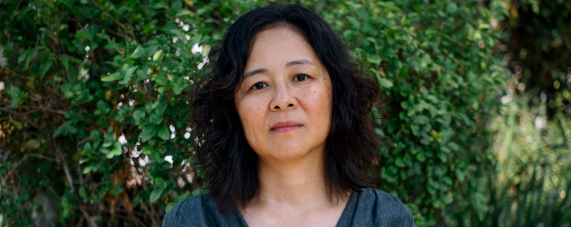 A person with medium-length, wavy dark hair and a neutral expression stands in front of a lush, green leafy background. Wearing a dark grey shirt, they gaze directly into the camera, embodying the composed demeanor often seen in Berkeley Journalism graduates.