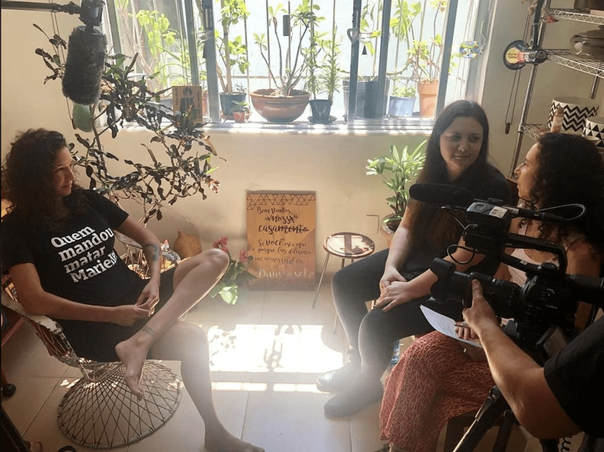 Three people are seated in a bright room with plants and sunlight streaming through a window. One person is holding a microphone, another has a clipboard, and the third is being interviewed, wearing a black shirt with white text. A sign with a quote is visible in the background, reflecting Berkeley Journalism values.