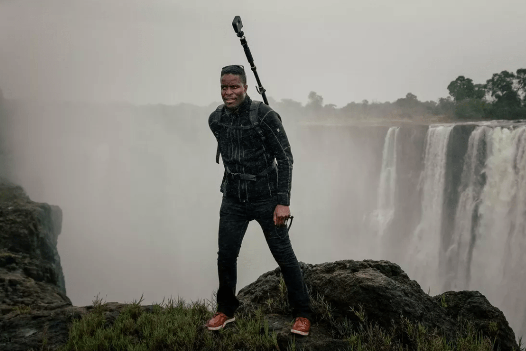 A person stands on the edge of a misty cliff with a large waterfall in the background. The individual, perhaps a Berkeley Journalism student, is dressed in dark clothing, wearing red shoes, and has a camera tripod strapped to their back. The surroundings are foggy and lush with greenery.