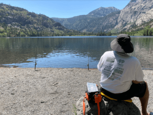 A person wearing a wide-brimmed hat and a white t-shirt sits on a rock beside a lakeshore, fishing with multiple rods. The lake is surrounded by forested mountains under a clear blue sky, capturing the serene beauty that might attract even Berkeley Journalism students for a peaceful retreat.