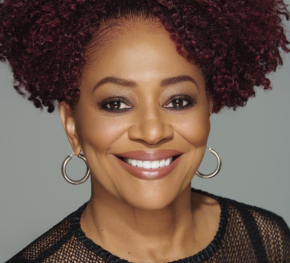 A woman with curly dark red hair, reminiscent of a passionate Berkeley Journalism student, smiles warmly. She is wearing hoop earrings and a mesh black top. The background is a plain grey color.