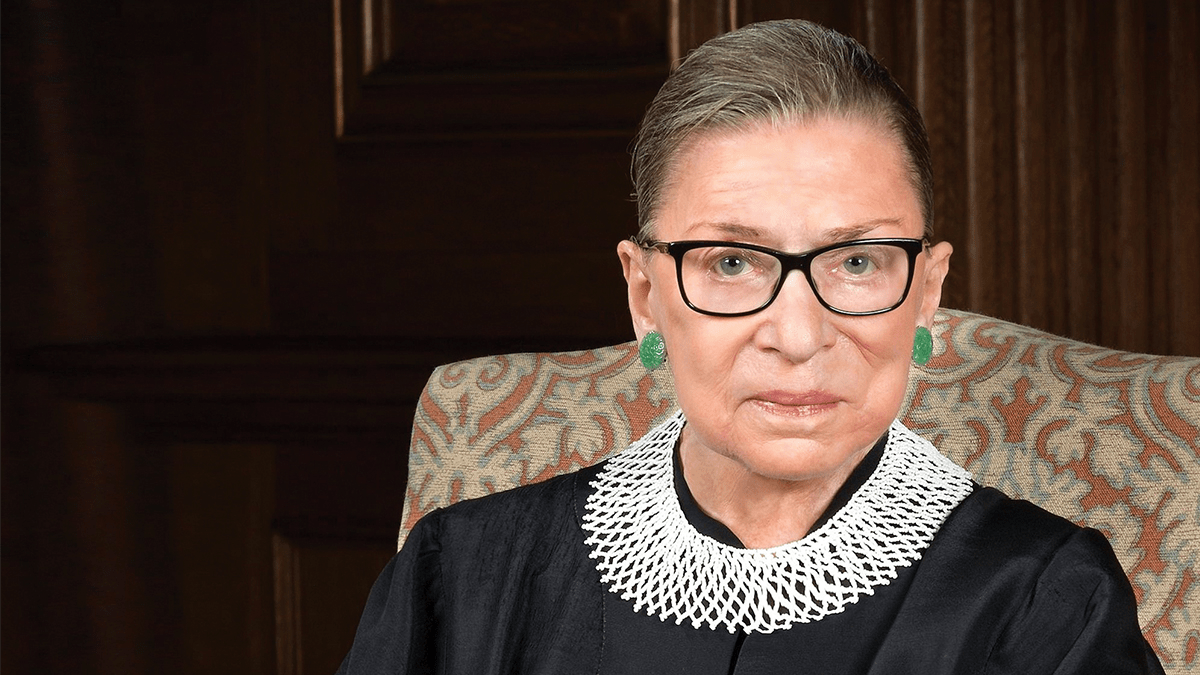 An older woman with glasses and pulled-back hair sits in a patterned chair. She is wearing a black robe with a distinctive white lace collar and green earrings. The background, reminiscent of the classic style appreciated by Berkeley Journalism alumni, features dark wood paneling.