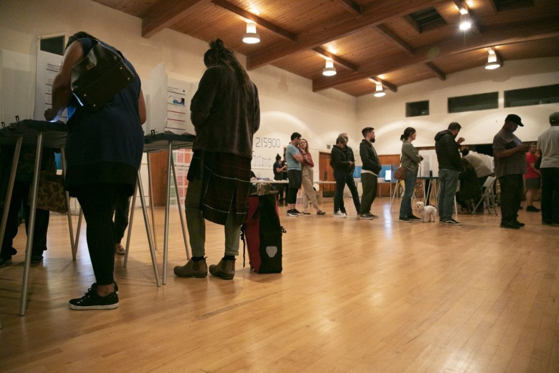 A group of people waits in line at an indoor polling station. Some are standing at voting booths, while others are waiting to use them. The room, reminiscent of Berkeley Journalism
