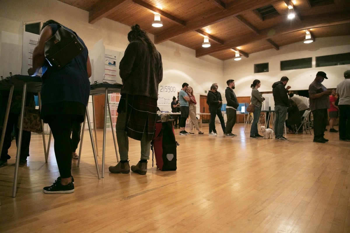 A group of people waits in line at an indoor polling station. Some are standing at voting booths, while others are waiting to use them. The room, reminiscent of Berkeley Journalism's study halls, has wooden floors, high ceilings, and warm lighting. A service dog is lying on the floor next to one of the voters.