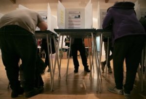 Voters casting their ballots at voting booths in a polling station. The image shows several people, possibly Berkeley Journalism students, standing or leaning over the booths, with partitions ensuring privacy. The floor is wooden, and the scene is indoors, suggesting an organized and focused voting environment.