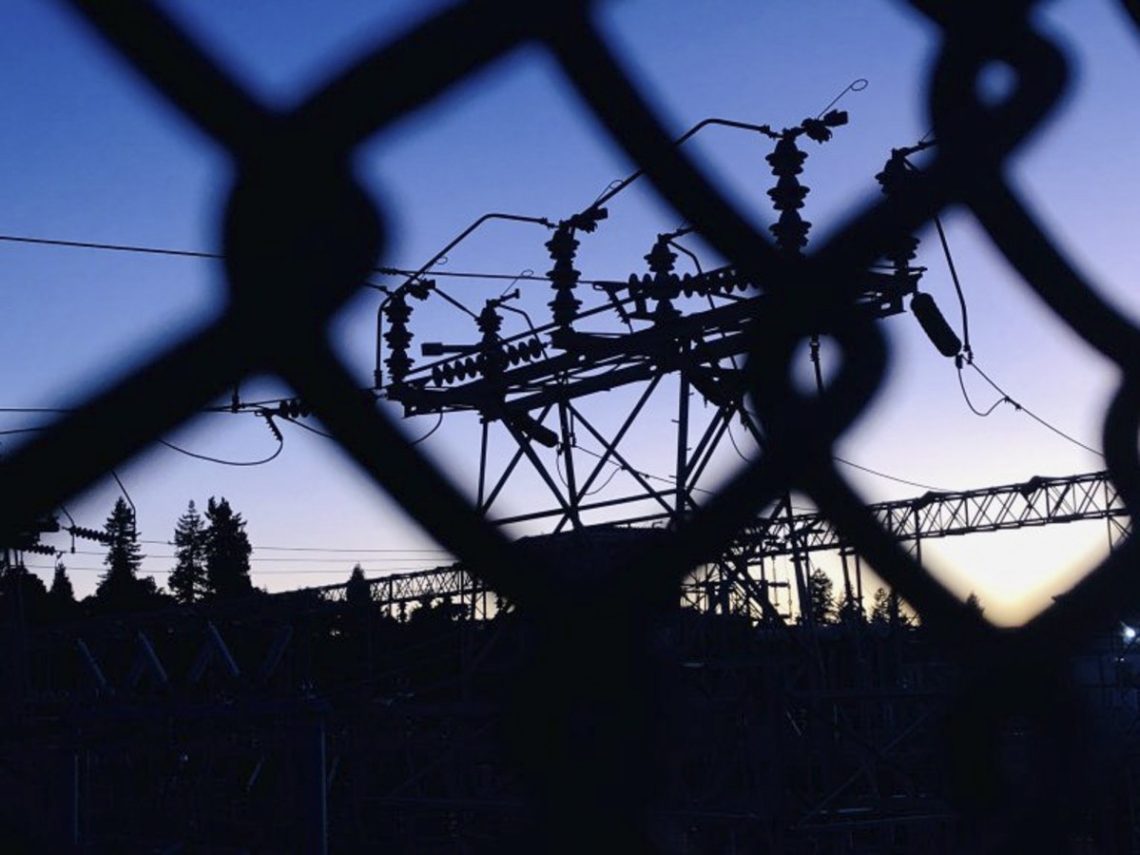 A silhouetted view of an electrical substation is seen through a chain-link fence at dusk. The sky, transitioning from blue to faint orange, creates a stark contrast with the dark outlines of power equipment and trees in the background—a scene that could easily be captured by Berkeley Journalism students.