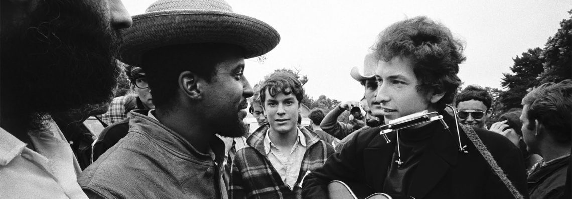 A diverse group of people gathers outdoors, with the focus on a person wearing a harmonica holder and holding a guitar. Another individual sporting a straw hat is engaged in conversation with them. The mood appears lively and sociable, resembling scenes often captured by Berkeley Journalism students.