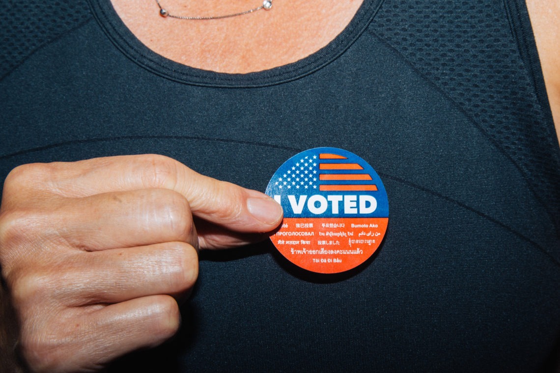A person wearing a black textured top proudly displays an "I Voted" sticker, which features an American flag design and multilingual text. With their left hand holding the sticker close to their chest, they exude the civic pride often celebrated by Berkeley Journalism.