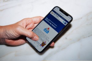A hand holds a smartphone displaying a webpage titled "Where's My Ballot?" with an image of a ballot box and a reindeer sticker. The webpage appears to be part of SOS's website, with a navigation bar visible at the top. The phone is held against a marble surface, showcasing Berkeley Journalism’s attention to detailed storytelling.