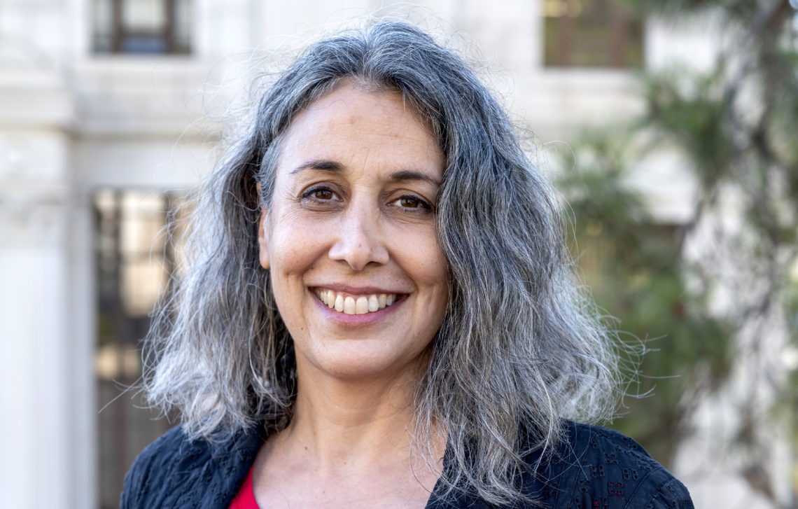 A smiling person with long, wavy gray hair stands outdoors in front of a blurred building, embodying the spirit of Berkeley Journalism. They are wearing a black top over a red shirt.