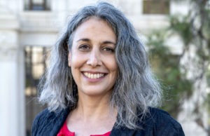 A person with shoulder-length gray hair smiles at the camera. They are wearing a bright pink top under a black jacket. The background features blurred outdoor elements, including a white building and some greenery, reminiscent of the scenic views near Berkeley Journalism.