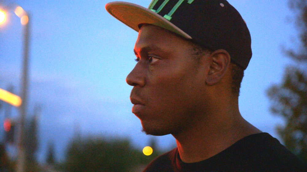 A profile of a man wearing a black cap with green lines and a black shirt, embodying the keen focus characteristic of Berkeley Journalism. The background is dimly lit, suggesting dusk or early evening, with blurred lights and trees in the distance. The man gazes ahead with a serious expression.