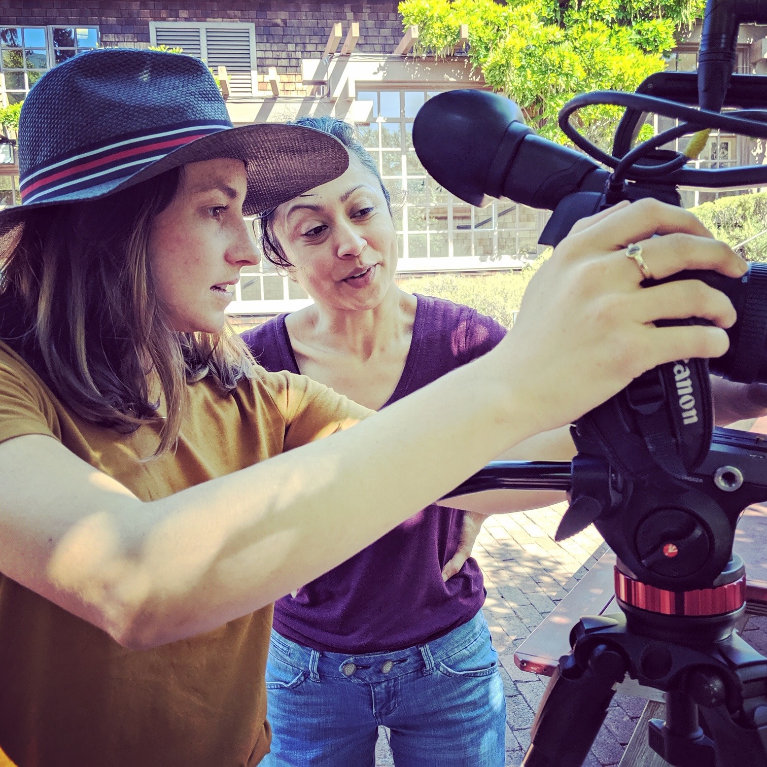 Two women from Berkeley Journalism are working together on an outdoor film set. One woman, wearing a hat, is adjusting the settings on a camera mounted on a tripod, while the other woman, standing beside her, is observing and discussing the setup. They are in a sunny courtyard with trees and buildings in the background.