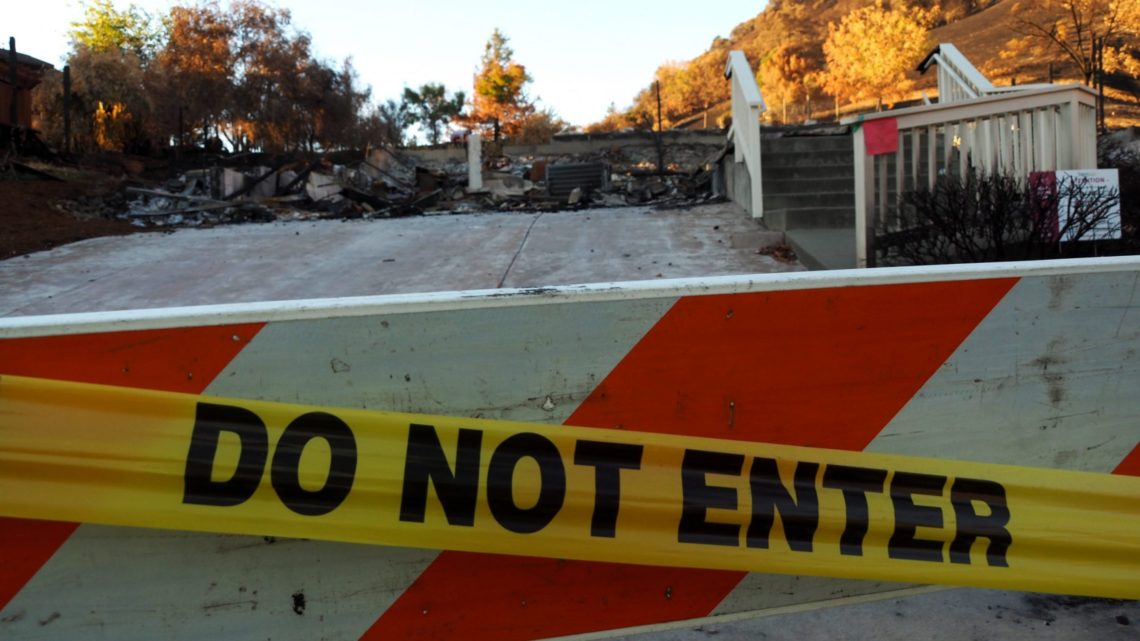 A "Do Not Enter" barricade tape stretches across the foreground, blocking access to a driveway leading to the remnants of a burned house. The area beyond the tape shows charred debris and the remains of a staircase. Captured by Berkeley Journalism, trees in the background display autumn colors.