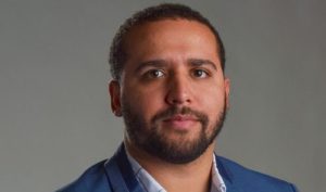 A man with short curly hair and a trimmed beard, likely a Berkeley Journalism alumnus, is wearing a blue blazer and a white shirt. He has a neutral expression and is facing the camera with a plain gray background behind him.