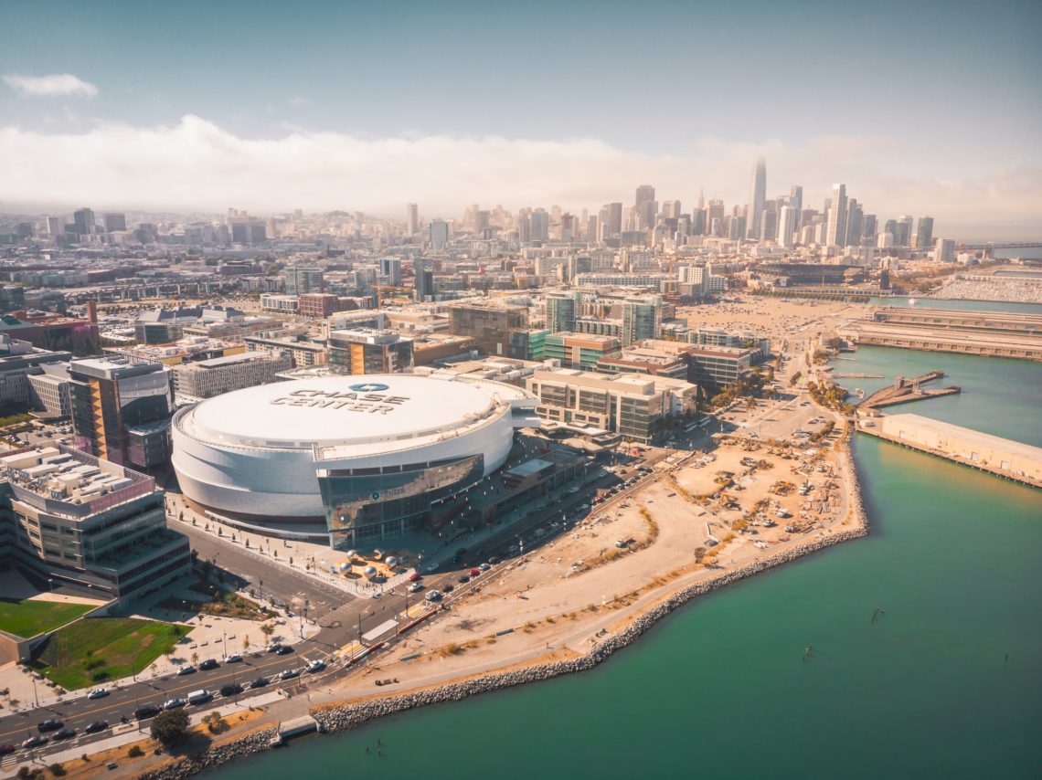 Aerial view of a coastal cityscape featuring the Chase Center, a large, white-domed arena. Surrounding the arena are buildings and infrastructure, with water bodies on two sides. The city skyline is visible in the background, partially shrouded in haze—a tableau that could captivate even Berkeley Journalism students.