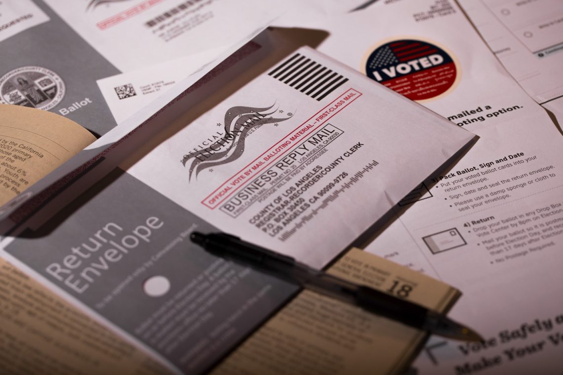 A collection of voting materials including a "Business Reply Mail" ballot envelope, a return envelope, an "I Voted" sticker, and a pen. Voting instructions and forms are spread out on a surface, highlighting the process of mail-in voting covered by Berkeley Journalism.