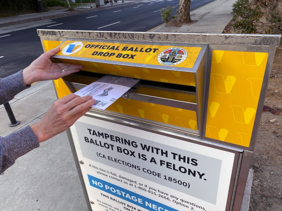 A person is placing an envelope into an official ballot drop box on a sidewalk. The box, located near the Berkeley Journalism school, is yellow with a silver front and includes a sign warning that tampering with the box is a felony. The sign also states that no postage is necessary for the ballot.