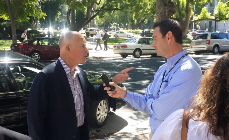 Two men are engaged in conversation on a sunny street lined with trees and parked cars. The man on the left, in a dark suit, is pointing while talking to the man on the right, who is holding a microphone marked with 