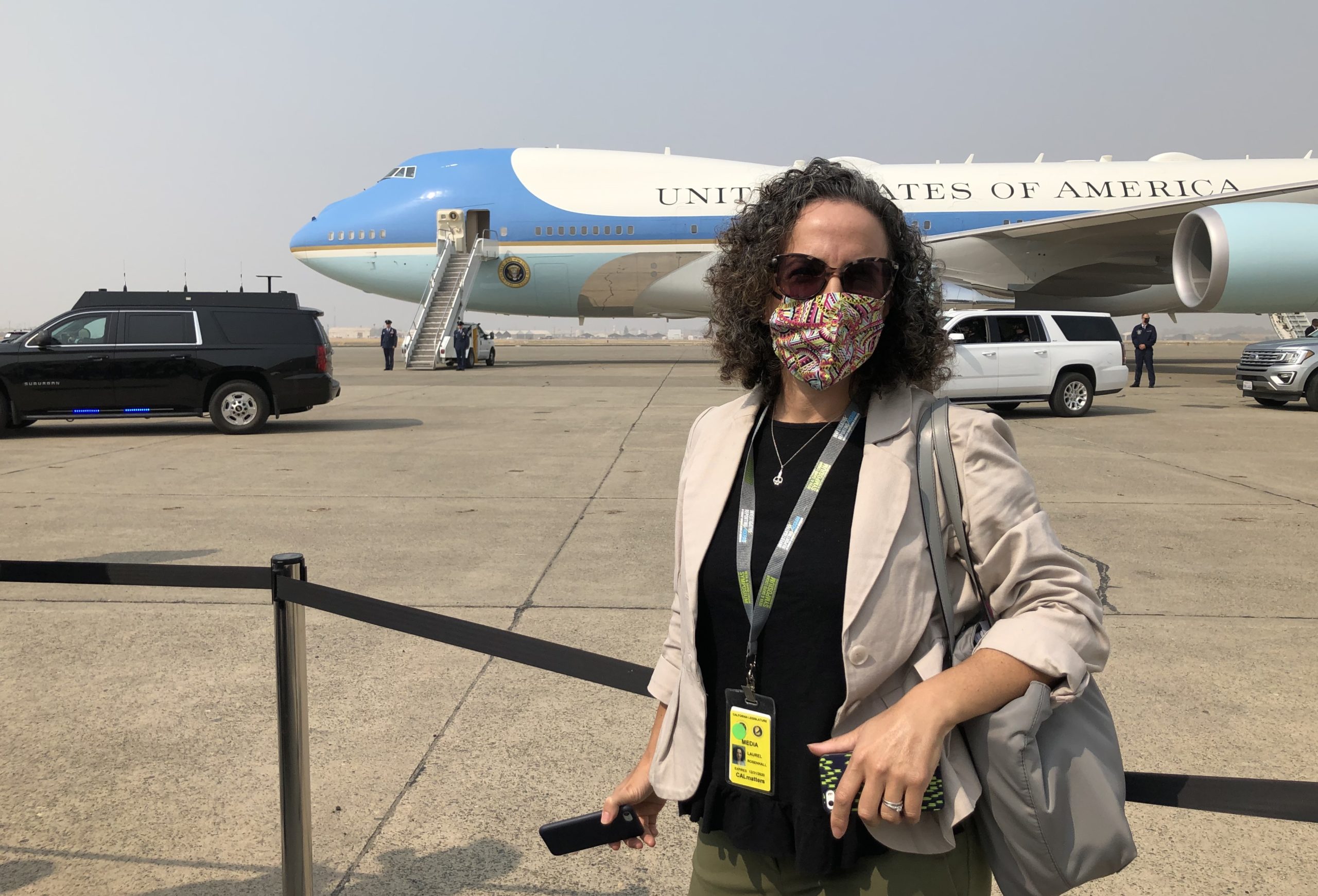 A woman with curly hair, wearing a floral mask and holding a phone, stands in front of Air Force One. Dressed in a light blazer and green pants, with a lanyard around her neck and a tote bag on her shoulder, she exudes the poise of Berkeley Journalism. Security vehicles are visible in the background.